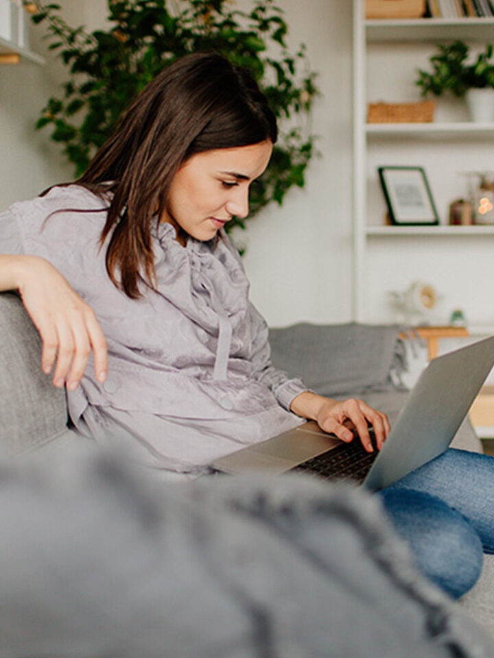Frau sitzt auf einem Sofa, schaut auf einen Laptop und tippt darauf, im Hintergrund sind eine Pflanze und Regale zu sehen.