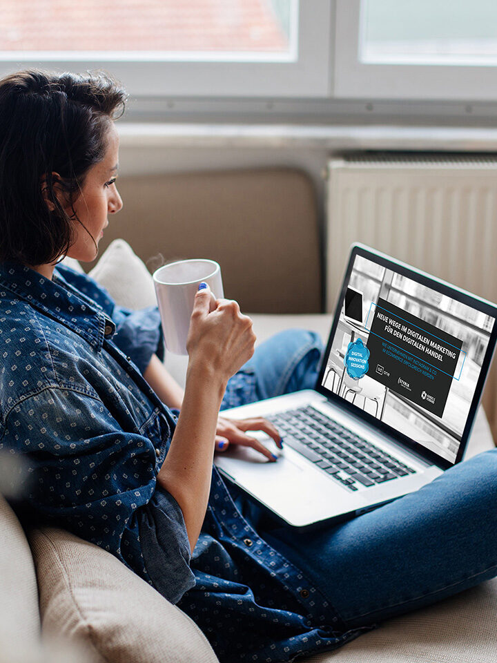 Frau sitzt auf einem Sofa, hält eine Tasse in der Hand und benutzt einen Laptop, auf dessen Bildschirm eine digitale Marketing-Webseite angezeigt wird.