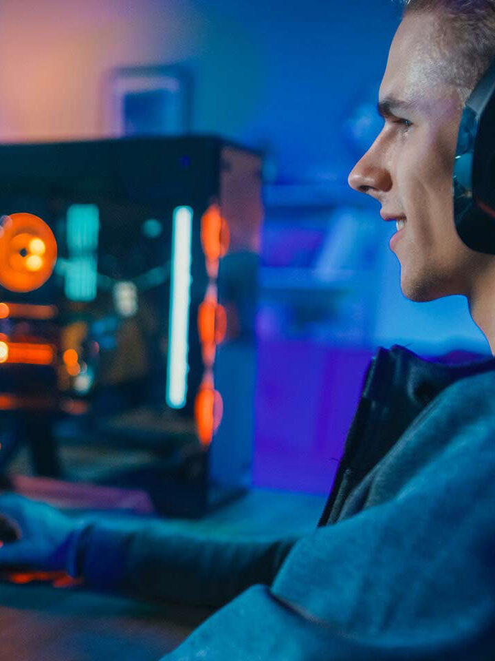 Ein junger Mann mit Kopfhörern sitzt an einem Schreibtisch und spielt in einem schwach beleuchteten Raum an einem Computer mit RGB-Beleuchtung.