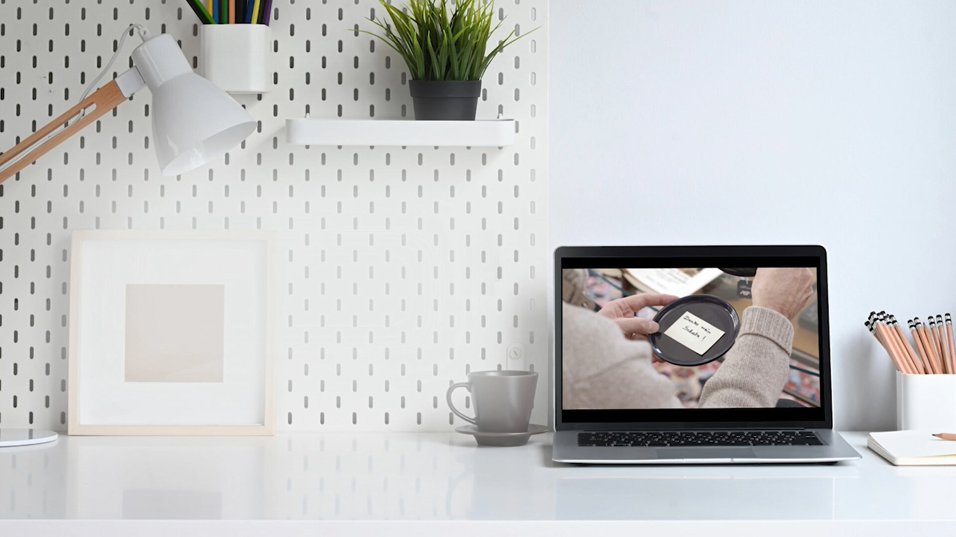 Ein Laptop auf einem weißen Schreibtisch zeigt ein Video, in der Nähe sind Büromaterial und Pflanzen aufgestellt.