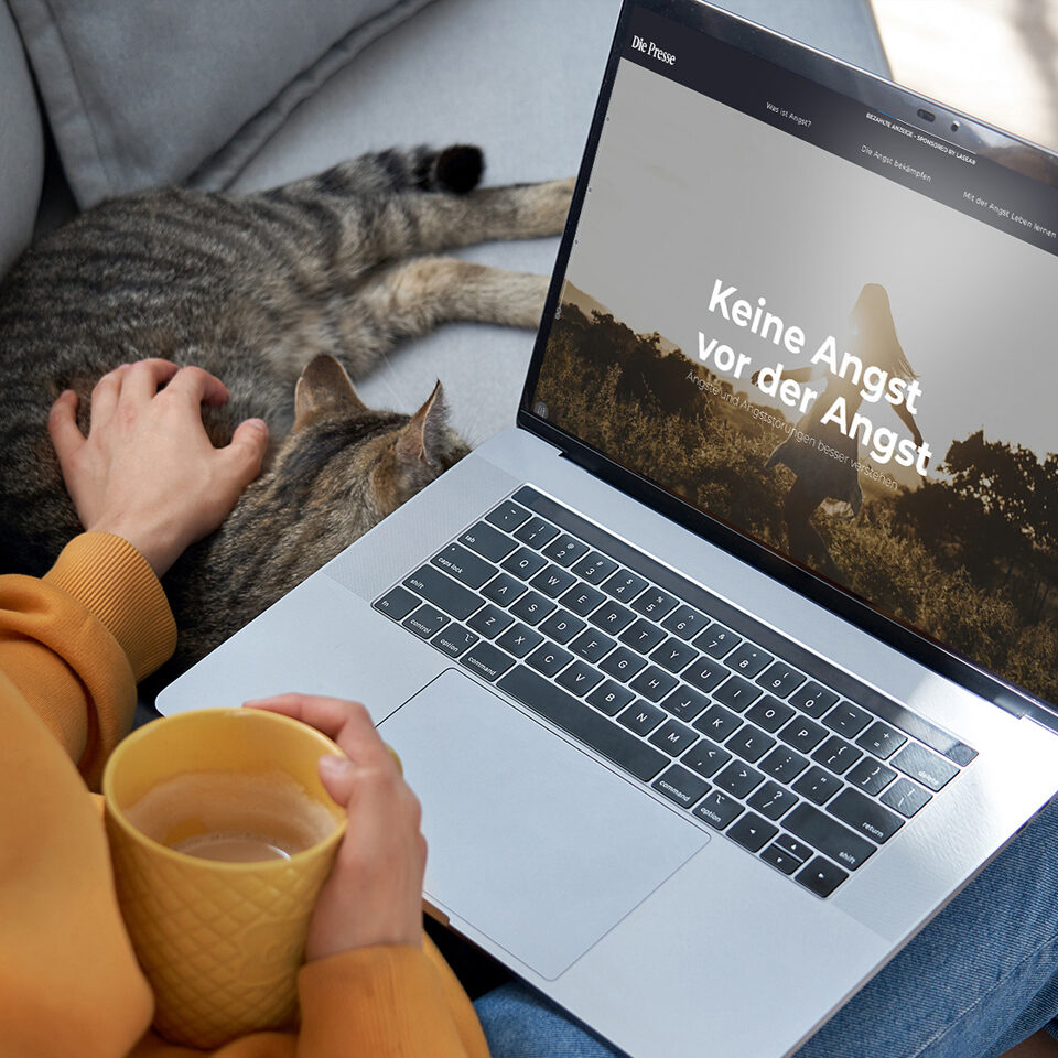Eine Person sitzt mit einer Katze auf einer Couch, hält eine gelbe Tasse in der Hand und benutzt einen Laptop, auf dem das Dossier 