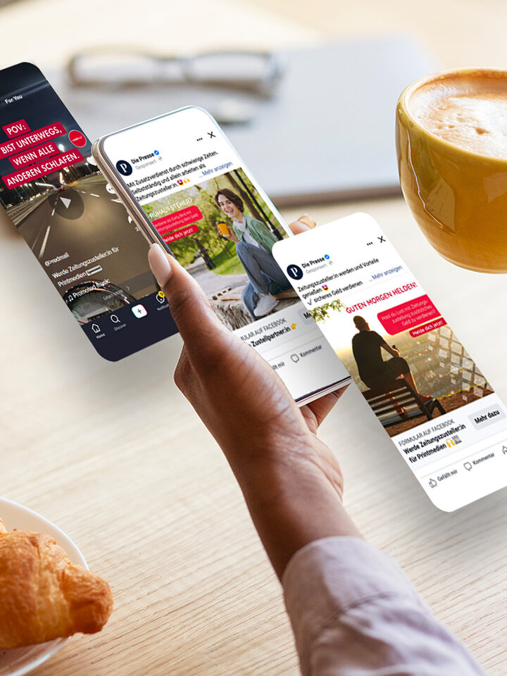 Eine Person hält über einem Tisch mit einem Croissant und einer Tasse Kaffee ein Smartphone, auf denen verschiedene Social-Media-Posts angezeigt werden, die die Wirkung von Performance Marketing veranschaulichen.