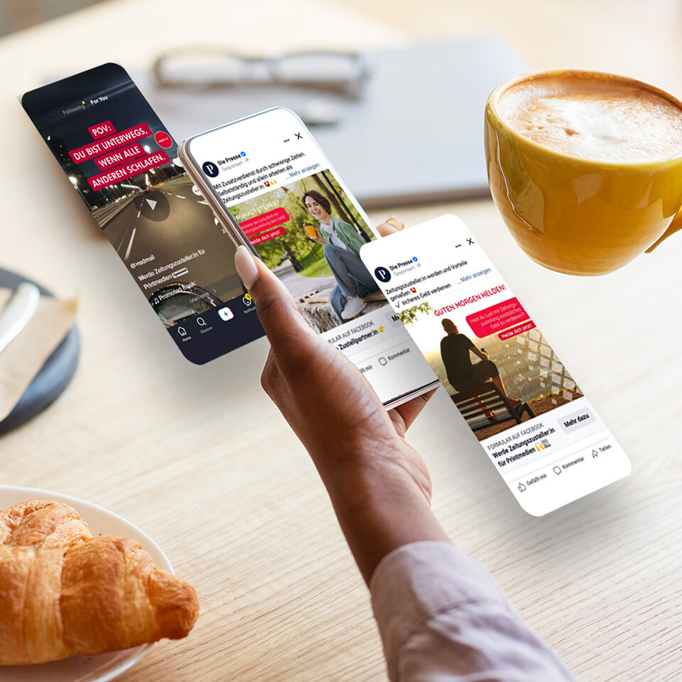 Eine Person hält über einem Tisch mit einem Croissant und einer Tasse Kaffee ein Smartphone, auf denen verschiedene Social-Media-Posts angezeigt werden, die die Wirkung von Performance Marketing veranschaulichen.