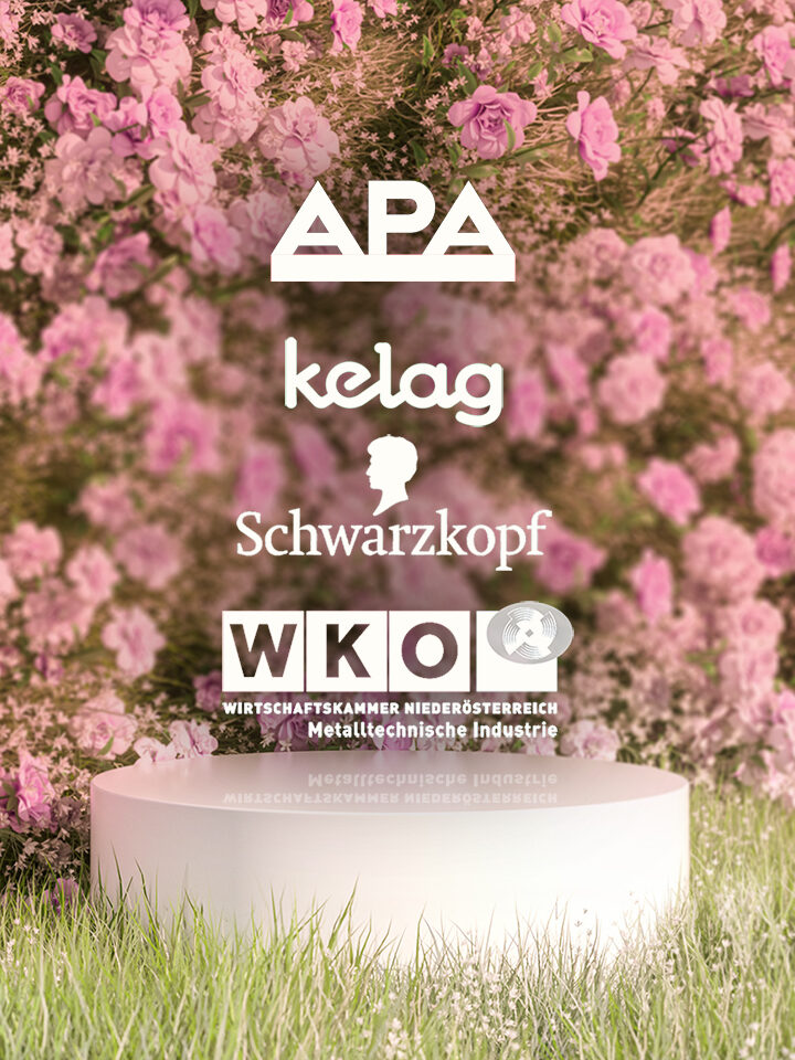Ein weißes Podest steht auf einer Wiese vor dichten rosafarbenen Blumen, darüber fünf Sponsorenlogos - darunter APA, Kelag, Schwarzkopf Professional und WKO