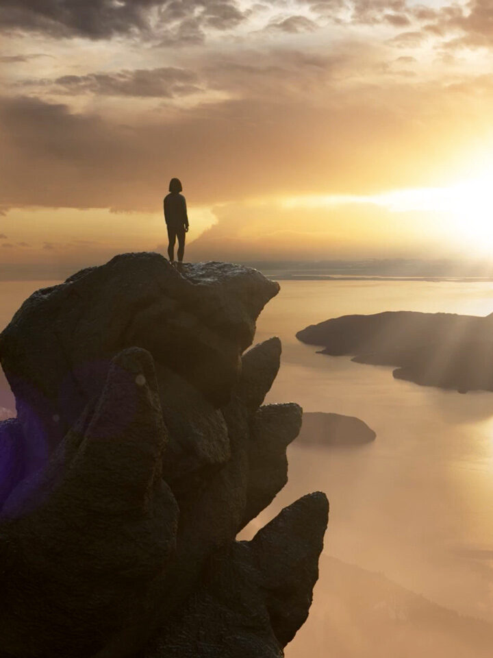 Eine Person steht auf einer felsigen Klippe und überblickt ein ruhiges Gewässer mit Inseln, während die Sonne hinter Wolken am Horizont untergeht oder aufgeht.