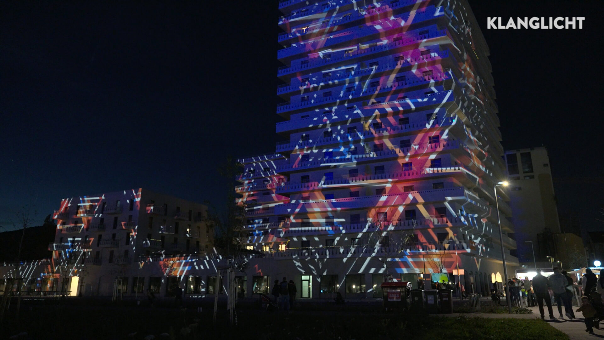 Auf der Fassade eines hohen Gebäudes werden nachts farbenfrohe geometrische Lichtprojektionen angezeigt, die im Rahmen einer Videoproduktion erstellt wurden. Am Fuße des Gebäudes sind Menschen und Bäume zu sehen.