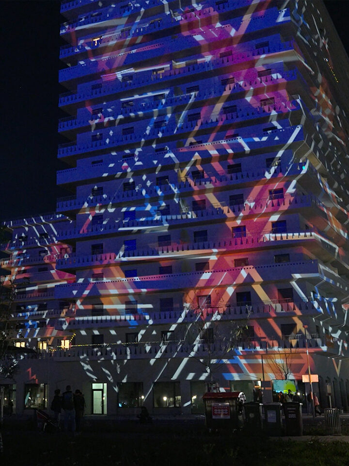 Auf der Fassade eines hohen Gebäudes werden nachts farbenfrohe geometrische Lichtprojektionen angezeigt, die im Rahmen einer Videoproduktion erstellt wurden. Am Fuße des Gebäudes sind Menschen und Bäume zu sehen.