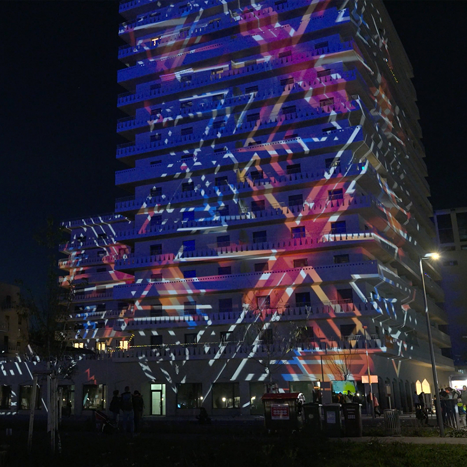 Auf der Fassade eines hohen Gebäudes werden nachts farbenfrohe geometrische Lichtprojektionen angezeigt, die im Rahmen einer Videoproduktion erstellt wurden. Am Fuße des Gebäudes sind Menschen und Bäume zu sehen.