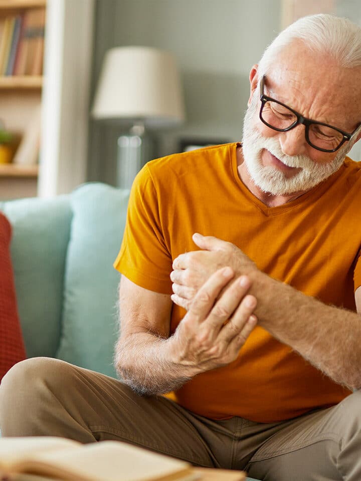 Ein älterer Mann mit Brille sitzt auf einem Sofa, hält mit einer Hand sein schmerzendes Handgelenk und zuckt zusammen. Vor ihm liegt ein offenes Buch.