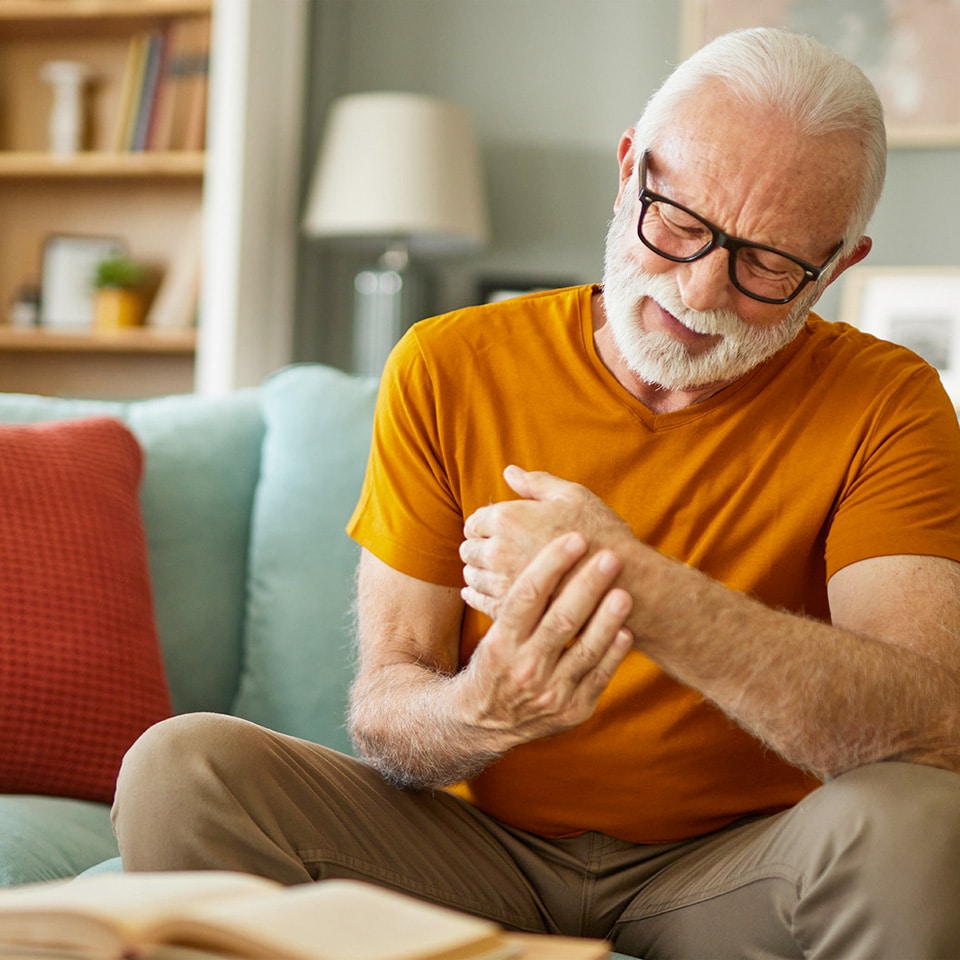 Ein älterer Mann mit Brille sitzt auf einem Sofa, hält mit einer Hand sein schmerzendes Handgelenk und zuckt zusammen. Vor ihm liegt ein offenes Buch.
