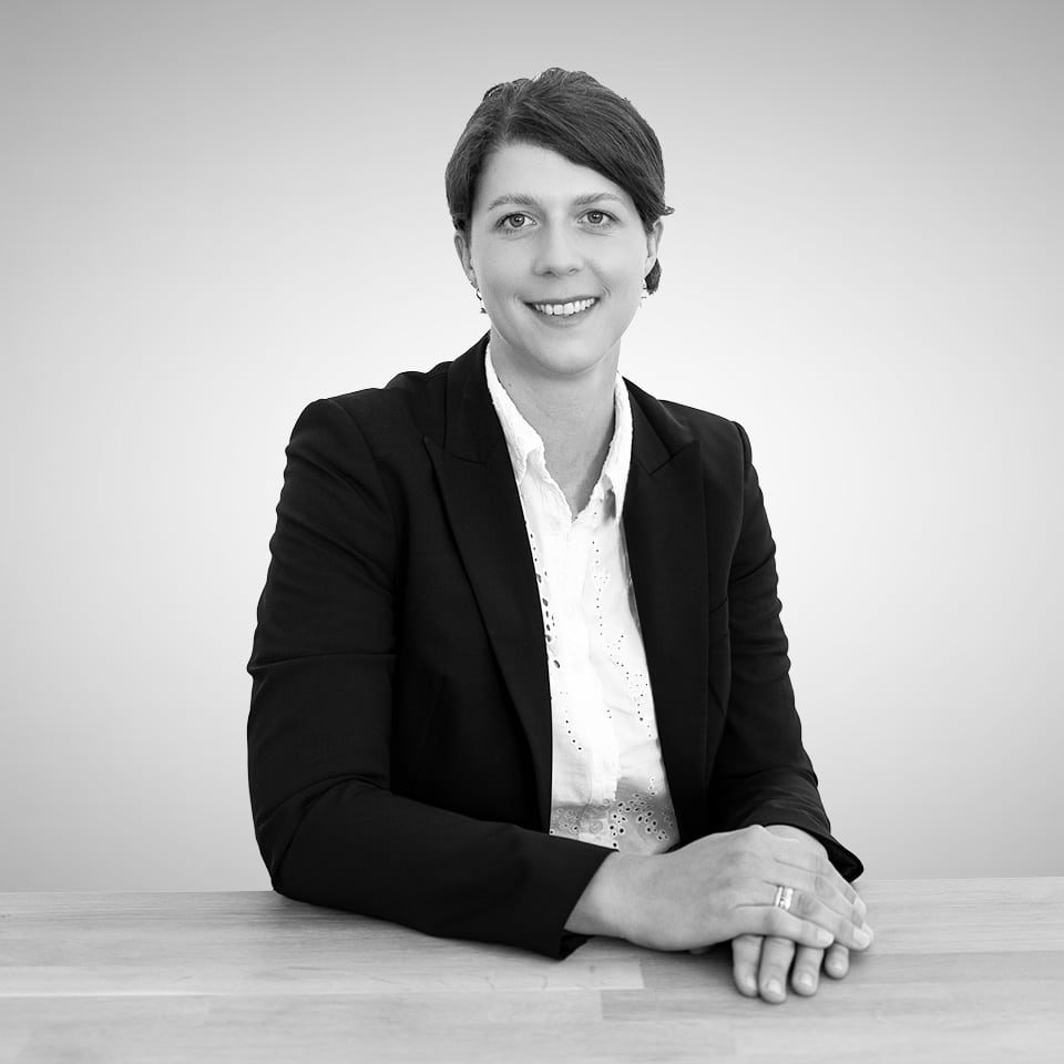 Eine Person in einem dunklen Blazer und weißem Hemd sitzt an einem Tisch, der Kamera zugewandt und lächelnd, die Hände auf die Tischplatte gestützt. Der Hintergrund ist einfarbig und hell.