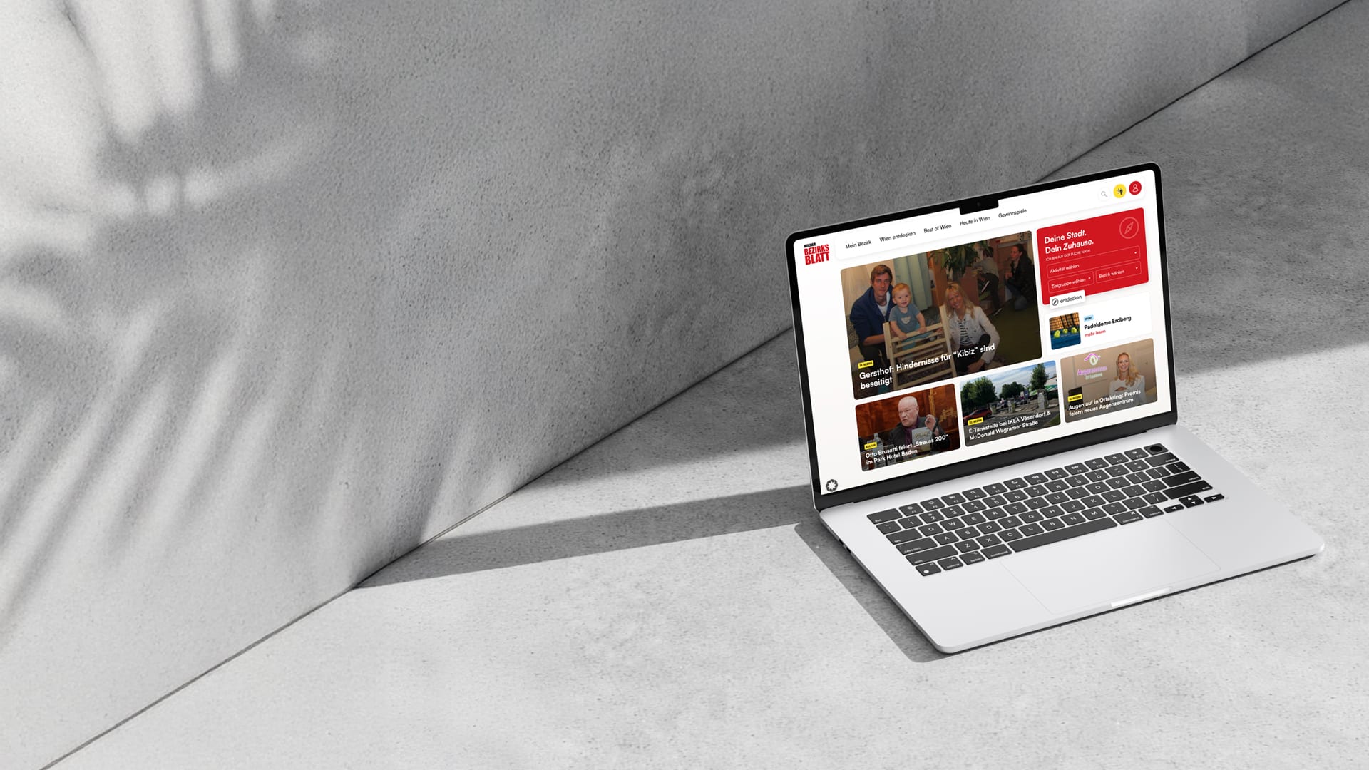Ein Laptop auf einer Betonfläche zeigt eine Nachrichten-Website an, wobei das Sonnenlicht und die Schatten der Blätter auf den Boden und die Wand fallen.