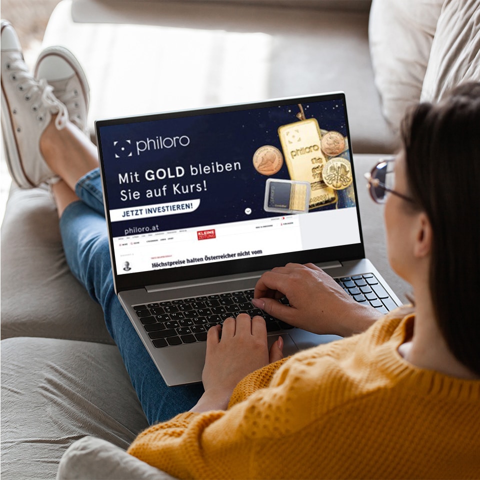 Eine Person, die auf einer Couch sitzt, benutzt einen Laptop, auf dem eine philoro-Website mit einer Werbung für deutsche Goldanlagen angezeigt wird.